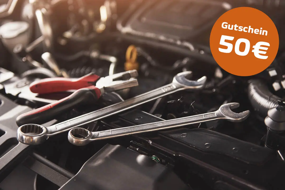 Nahaufnahme eines Motorraums: Auf dem Aggregat liegen zwei Schraubenschlüssel und eine Zange; rechts oben ein orangefarbener Kreis mit der Aufschrift „Gutschein 50 €“.
